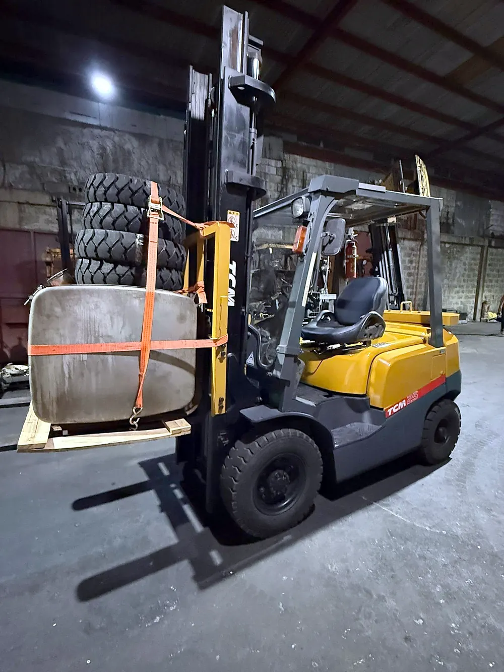 TCM 25 forklift carrying a strapped tank and stacked tyres in a warehouse